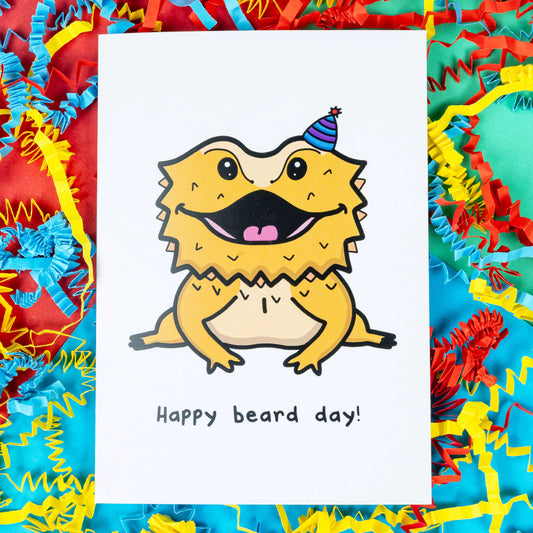 Happy Beard Day - Bearded Dragon A6 Birthday Card on a green, blue and red background with yellow, blue and red crinkled card confetti. The white card features a happy bearded dragon reptile wearing a purple stripe party hat, underneath in black reads Happy Beard Day!