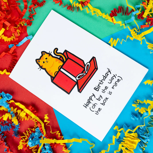 A white card with a ginger cat illustration coming out of a red present with text saying Happy Birthday! (Oh by the way, the box is mine). The a6 birthday card is laying on a red, blue and green background with red, yellow and blue crinkle card confetti.
