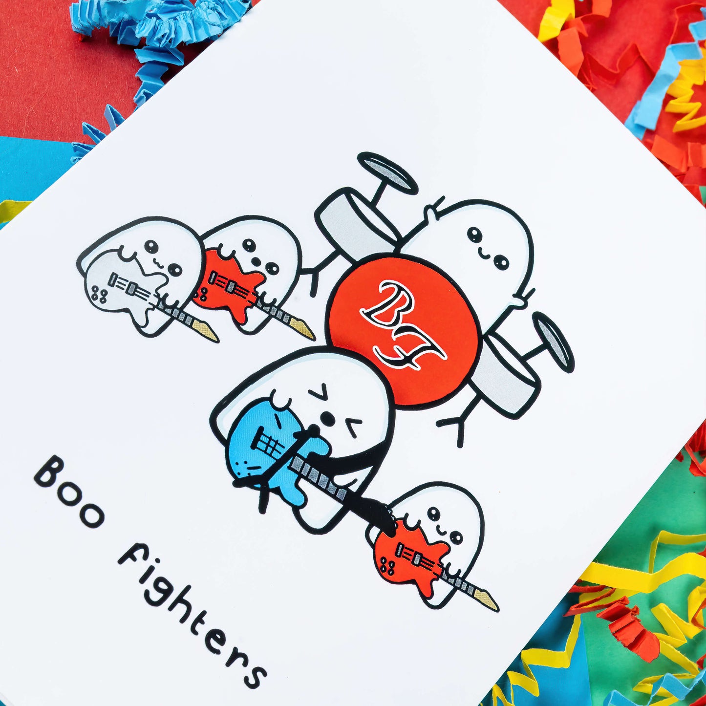 Close up of the Boo Fighters Birthday Card on a green, blue and red background with red, blue and yellow crinkle confetti card. A white card with a kawaii ghost band playing blue, white and red guitars and a red Boo Fighters drum kit, underneath reads Boo Fighters in black text.