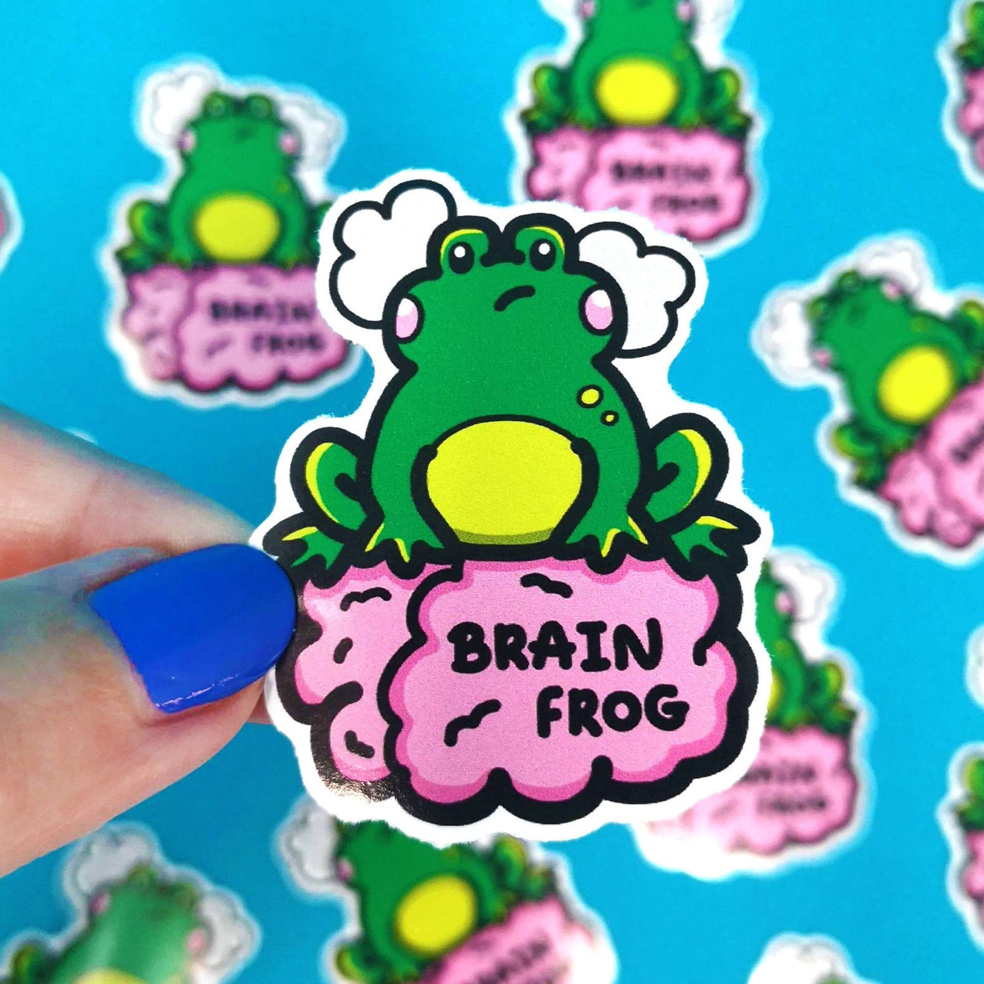 The Brain Frog Sticker - Brain Fog being held above multiple frog stickers by a hand with blue nail varnish over a blue background. The sticker is a confused frog with pink blush cheeks and two grey clouds above its head. Its sat on a pink brain with text that reads Brain Frog. The design was created to raise awareness for brain fog.