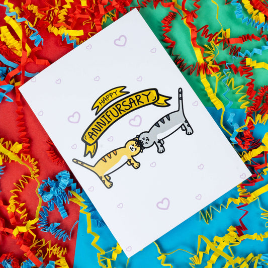 a white greeting card with light pink hearts dotted all over. 'Happy ANNIFURSARY' is written in a gold banner on the card with two cat illustrations underneath, one ginger and one grey. The cats are rubbing their heads together. The background of the photo is red, blue and green card with paper card decorations.