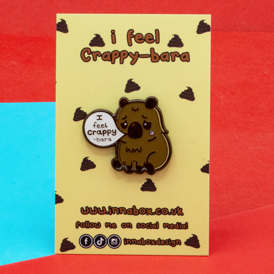 I Feel Crappy-Bara enamel pin badge by Innabox, displayed on a yellow backing card with the words “I feel Crappy-bara” and small poop illustrations. The kawaii-style capybara has a sad expression and speech bubble saying “I feel crappy-bara”. Disability-inspired design raising awareness for depression, chronic illness, and hidden disabilities.