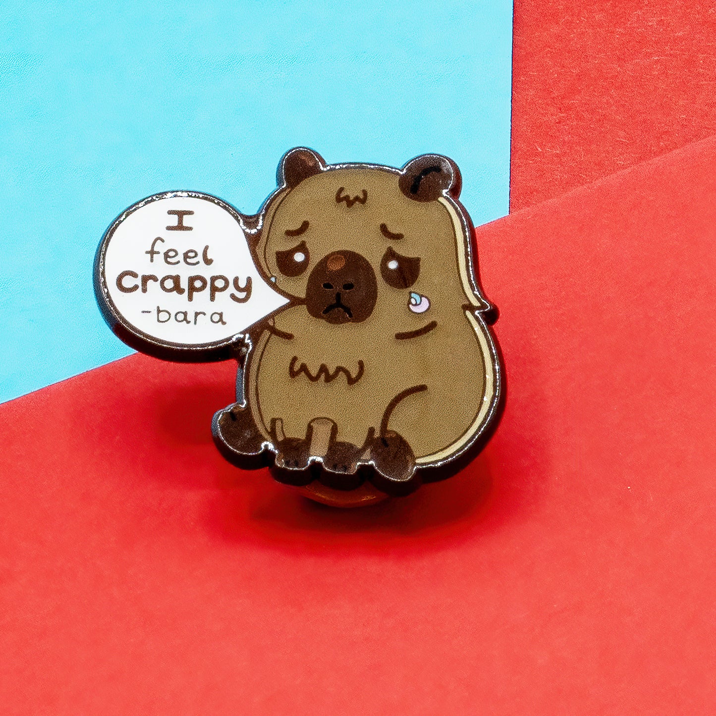 Innabox I Feel Crappy-Bara enamel pin badge on red and blue background. Features a sad capybara with tearful eyes and a speech bubble saying “I feel crappy-bara”. Disability-inspired pin for mental health, depression, and chronic illness awareness.
