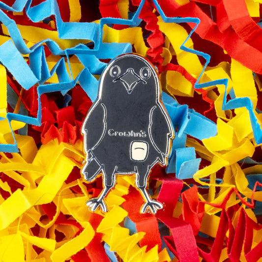 The Crowhn's Disease Enamel Pin - Crohn's Disease on a red, blue and yellow card confetti background. The black crow shape pin has a white stoma bag fitted and the text reading 'chrowhn's' across its chest. The design is raising awareness for crohn's disease.