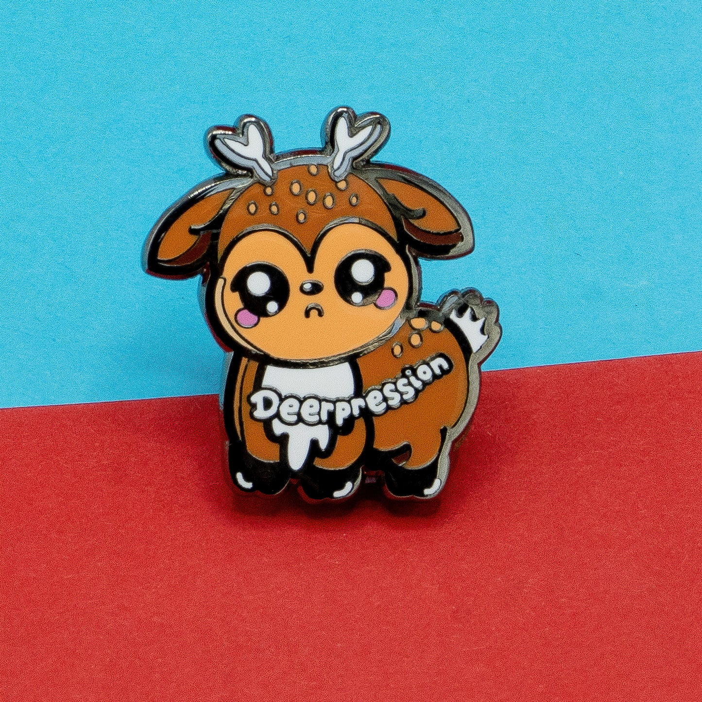 Close-up of the Innabox Deerpression 2.0 enamel pin featuring a cute sad-eyed deer character with antlers and blushing cheeks. The word "Deerpression" is written across its side. Disability-inspired design created to support awareness of depression, chronic illness, and hidden disabilities.