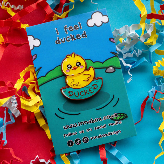 Innabox I Feel Ducked enamel pin displayed on branded backing card with blue sky and pond illustration. The 30mm hard enamel pin shows a kawaii yellow duck with glossy finish and uplifting awareness message, photographed on bright confetti background.