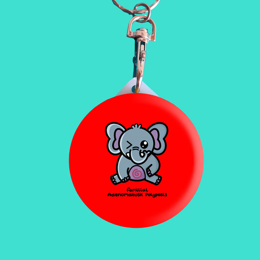 a red circular keyring with a cute elephant illustration on the front. The elephant is sat on it's back legs and has one eye shut. The elephant has a red spiral on it's tummy and is holding it's tummy. 'familial adenomatusk polyposis' is written underneath. The background of the photo is blue.