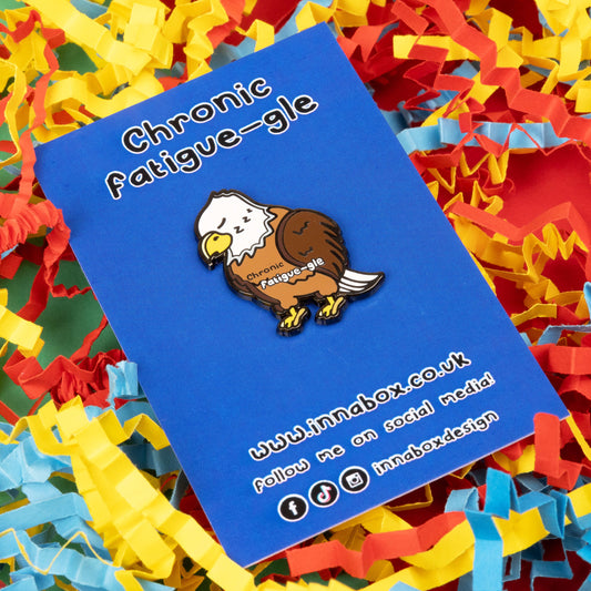 The Chronic Fatigue-le Enamel Pin - Chronic Fatigue on it's blue backing card with Innabox social media handles on a red, blue and yellow card confetti background. The enamel pin is a sleeping tired brown and white eagle with a yellow beak and feet, across the middle in black text reads 'chronic' and fatigue-gle' in white text. The design was created to raise awareness for chronic fatigue / ME / CFS.