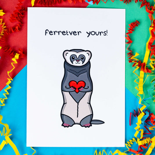 The Ferretver Yours Valentines Day Love Card on a red, blue and green background with red, yellow and blue crinkle card confetti. A white a6 greetings card with a drawing of a happy ferret holding a red heart saying ferreter yours on the front. 