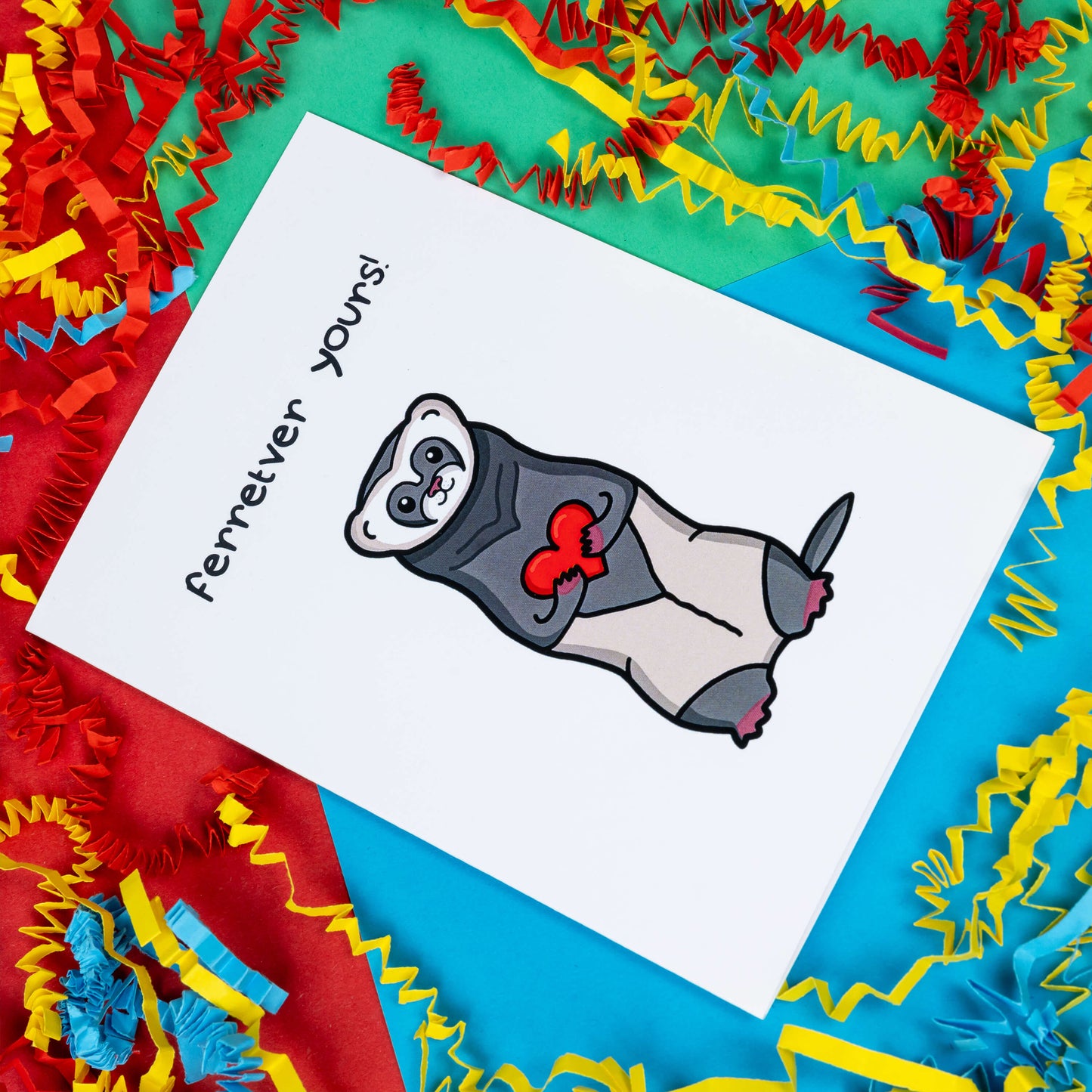 The Ferretver Yours Valentines Day Love Card on a red, blue and green background with red, yellow and blue crinkle card confetti. A white a6 greetings card with a drawing of a happy ferret holding a red heart saying ferreter yours on the front.