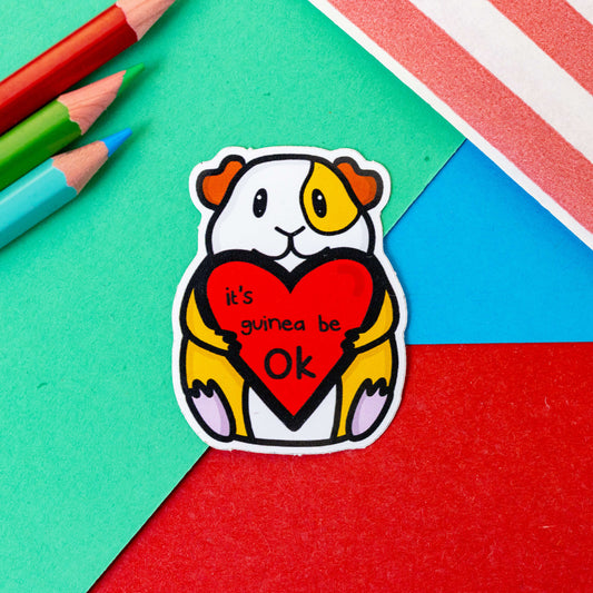 The It's Gonna Be OK Guinea Pig Sticker on a red, blue and green background with colouring pencils and red stripe candy bag. The guinea pig shape sticker is orange and white smiling sat down holding a red heart with black text reading 'it's guinea be ok'. The hand drawn design is a reminder to stay positive.