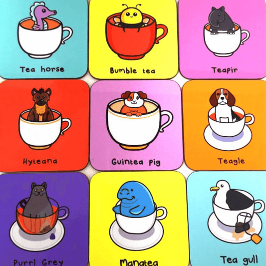 The Guinea Pig Tea Coaster amongst other innabox tea pun coasters. The pastel pink wooden coaster has a smiling brown and white guinea pig popping out of a white mug of tea, underneath in black reads 'guintea pig'.