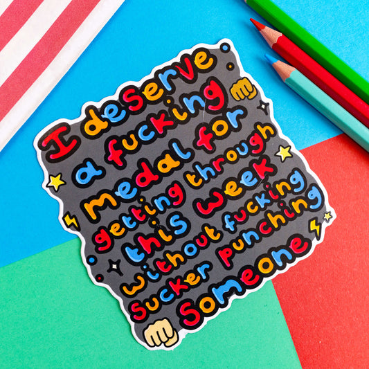 I Deserve A Medal Sticker of the words I deserve a fucking medal for getting through this week without fucking sucker punching someone' in bubble text with hand, lightning bolts, sparkles and star emojis. It is on a red, blue and green background with coloured pencils to one side and a stripy paper bag.