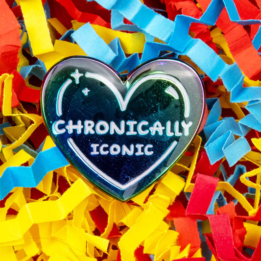 The Chronically Iconic Enamel Pin on a red, blue and yellow card confetti background. The anodised rainbow heart shaped pin has a white outline with sparkles and text reading 'chronically iconic'. The design was created to raise awareness for chronic illness and invisible illness.