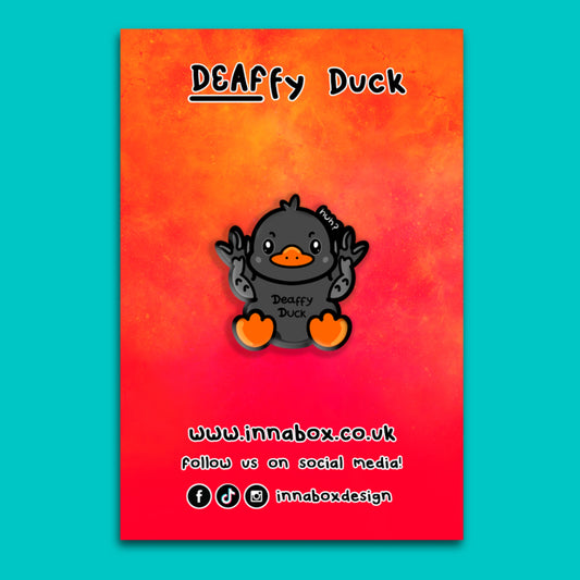 Illustrated backing card for the Deaffy Duck enamel pin by Innabox, featuring a cute black duck character using British Sign Language for deaf. The background fades from orange to pink with Innabox branding, website URL and social media icons. Disability inspired design created to uplift people with chronic illness, deafness and hidden disabilities.