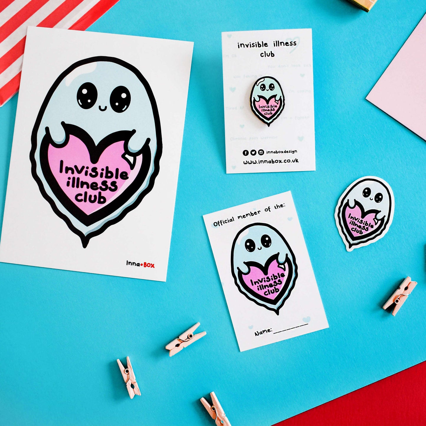 The Invisible Illness Club Sticker on a blue background with various other innabox products. The pastel blue smiling ghost sticker has big sparkly eyes holding a pastel pink heart with black text reading 'invisible illness club'. The hand drawn design is raising awareness for hidden disabilities.