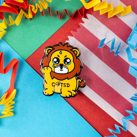 The Complex Post Troarmatic Stress Disorder Lion Enamel Pin - C-PTSD - Complex Post Traumatic Stress Disorder on a red, blue, green and yellow crinkle card confetti background. A yellow crying smiling and sweating male lion with its paw raised in the middle with 'C-PTSD' across its chest. The enamel pin design is raising awareness for complex post traumatic stress disorder.