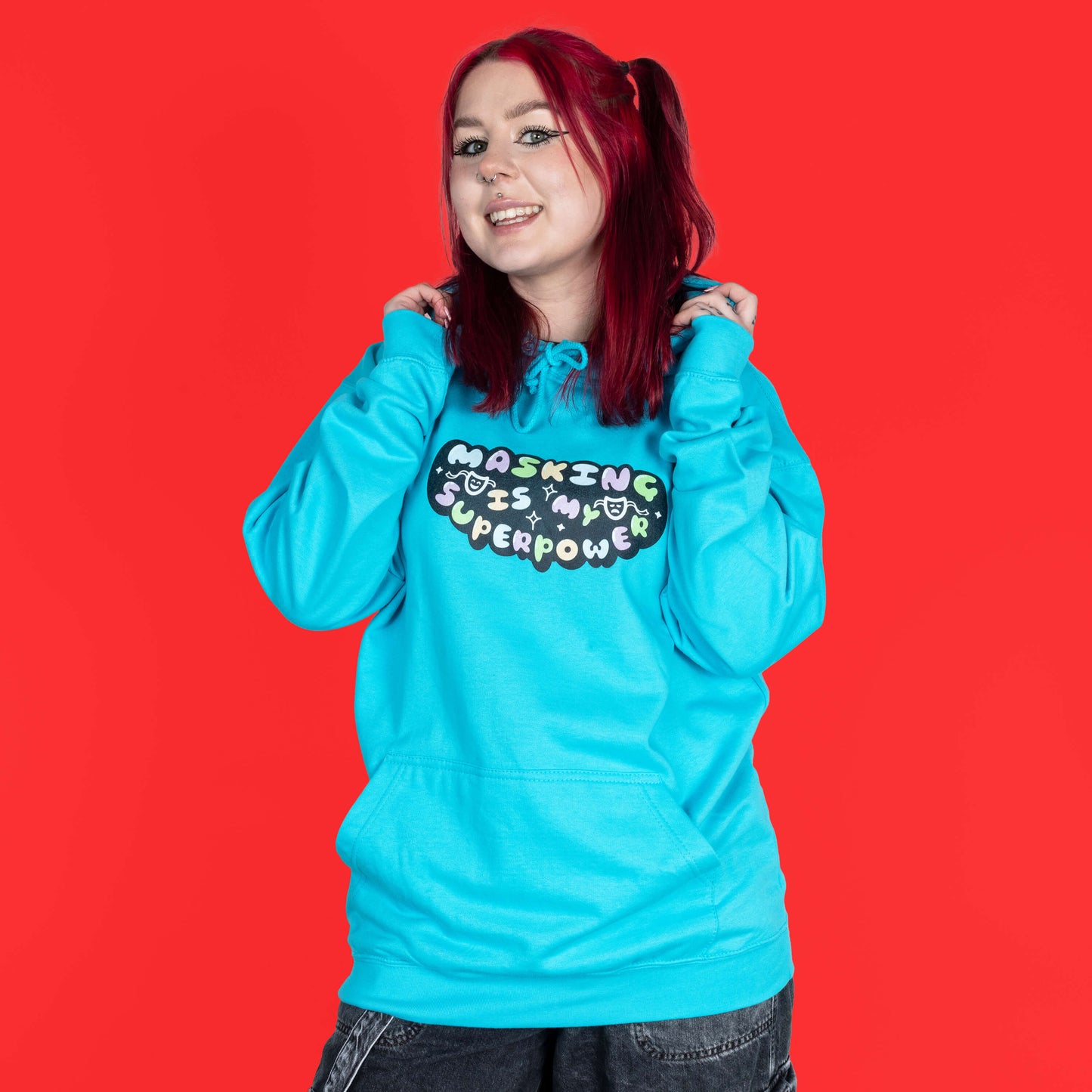 The Masking Is My Super Power Hoodie in Turquoise Surf modelled by Flo with red hair and black eyeliner on a red background. She is facing forward smiling holding the hood. The aqua blue hoodie features pastel rainbow bubble writing that reads 'masking is my superpower' with white sparkles, a happy and sad drama masks all on a black oval. Raising awareness for neurodivergent people with ADHD or Autism.