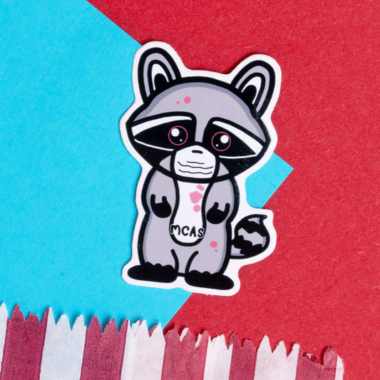 Innabox MCAS raccoon sticker featuring a cute, grey cartoon raccoon with a medical mask, symbolising Mast Cell Activation Syndrome (MCAS). The raccoon is designed with pink patches, big eyes, and the text 'MCAS' on its chest. The background is a mix of blue and red paper, enhancing the sticker's bold and uplifting design.