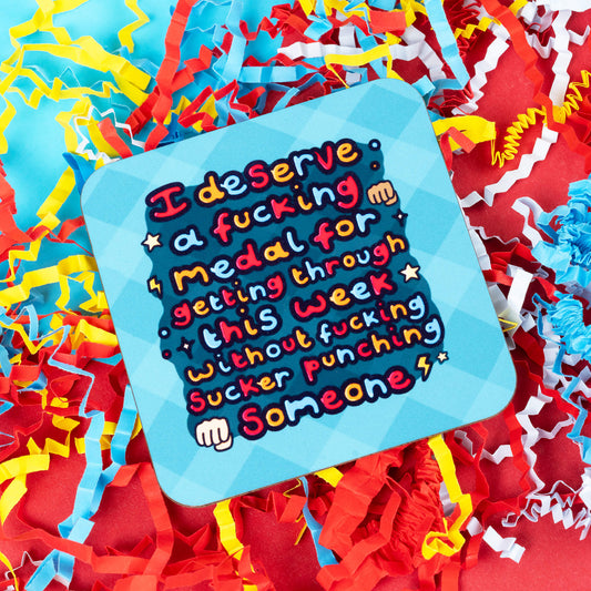 A colourful Innabox coaster with a bold hand-drawn quote that reads, "I deserve a fcking medal for getting through this week without fcking sucker punching someone", surrounded by red, yellow, and blue paper confetti. The coaster is square with rounded corners and sits on a bright blue background. Designed to support people with chronic illness and hidden disabilities.
