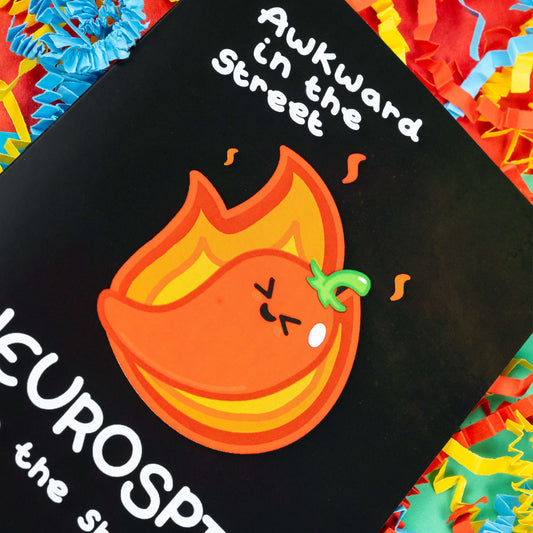 Close up of A black greeting card with a cute illustration of a red chilli pepper with scrunched up eyes and big smile. There is orange fire behind the chilli. 'Awkward in the street NEUROSPICY in the sheets!' is written on the card in white. The background of the photo is colourful card confetti. Design inspired by neurodivergent disorders.