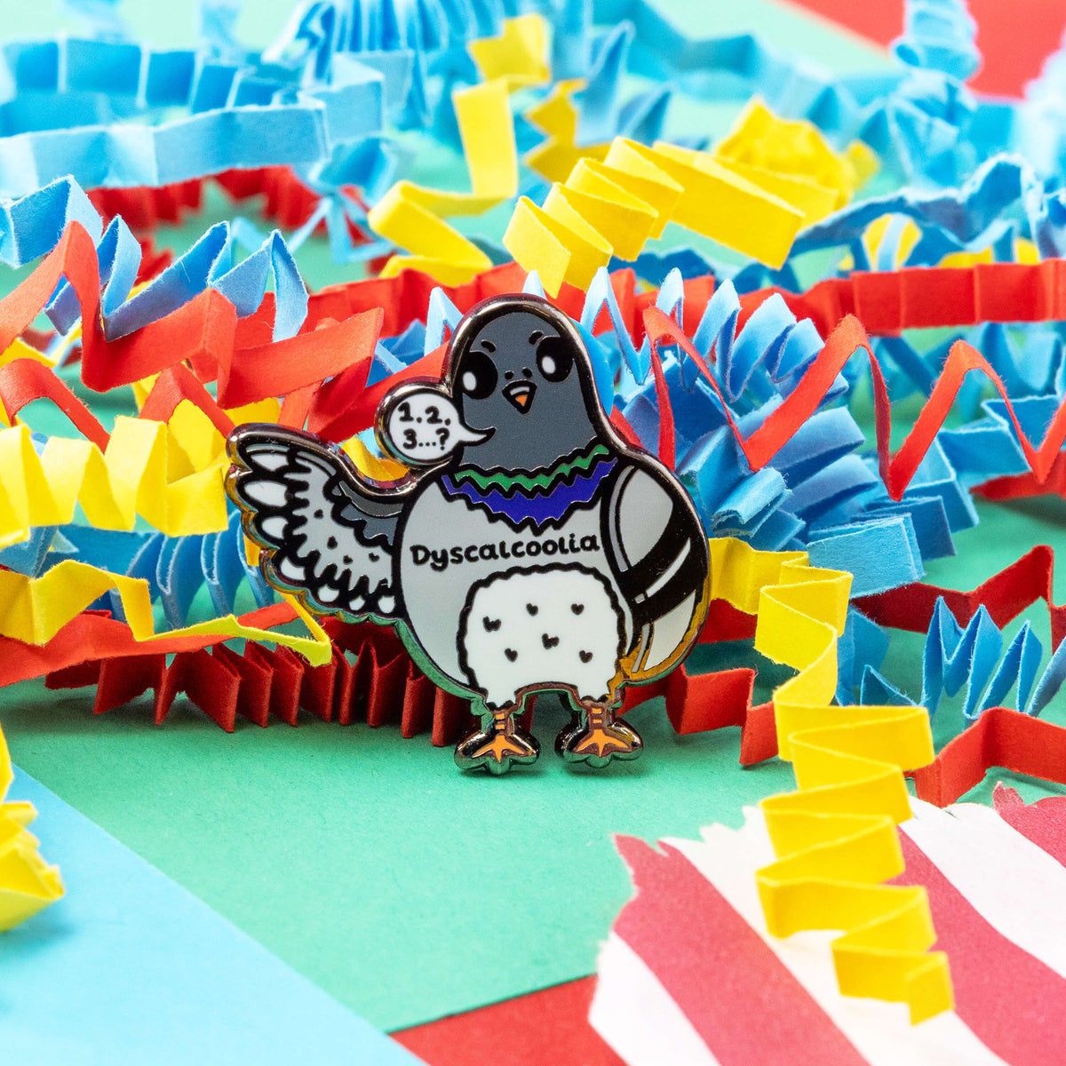 The Dyscalcoolia Pigeon Enamel Pin - Dyscalculia on a red, blue, green and yellow crinkle card confetti background. A Pigeon looking confused holding up a wing with a speech bubble full of numbers and a question mark across its chest reads 'dyscalcoolia'. The enamel pin is raising awareness for dyscalculia.