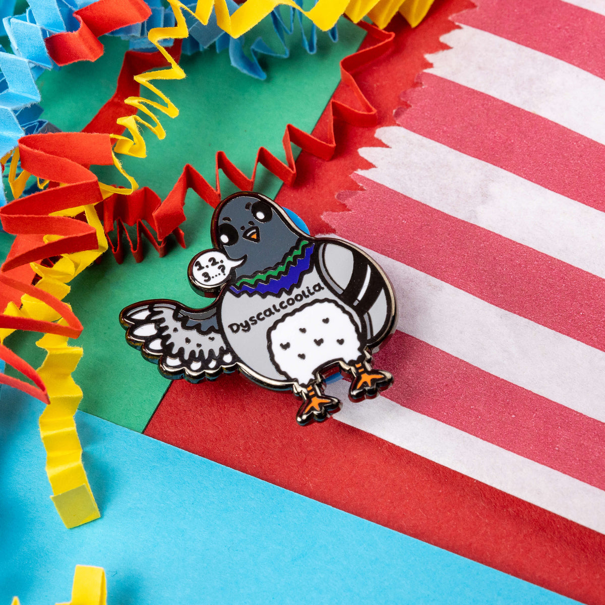 The Dyscalcoolia Pigeon Enamel Pin - Dyscalculia on a red, blue, green and yellow crinkle card confetti background. A Pigeon looking confused holding up a wing with a speech bubble full of numbers and a question mark across its chest reads 'dyscalcoolia'. The enamel pin is raising awareness for dyscalculia.