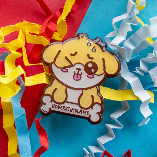Close up of Roverstimulated Dog enamel pin by Innabox on bright blue background with paper streamers. Glossy 30mm hard enamel pin featuring an overwhelmed yellow dog with sweat drops and “Roverstimulated” bone, raising awareness for hidden disabilities.