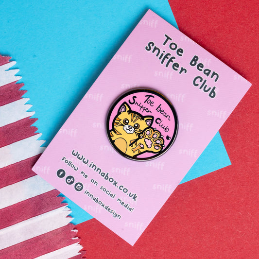 Innabox pin badge on a pink backing card with 'Toe Bean Sniffer Club' text. The badge features an orange cat with raised paw and pink toe beans, against a pink background. Innabox, known for its disability-inspired products, created this item to uplift individuals with chronic illnesses or hidden disabilities.