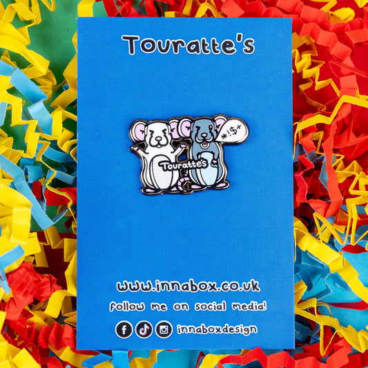 The Touratte's Rat Enamel Pin - Tourette's Syndrome on blue backing card laying on a red, blue and yellow paper background. The enamel pin is of two rats with pink ears and feet, the white rat on the left is raising its arms with its eyes closed and the grey rat on the right has a speech bubble with symbols in, across both rats reads 'touratte's'. The hand drawn design is raising awareness for tics and tourettes.