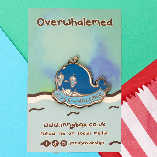 Innabox's Overwhalemed Glittery Whale Enamel Pin displayed on a pastel gradient backing card with a wavy sea design. The pin shows a sad blue whale with big, watery eyes and a glittery blue wave beneath it, with "Overwhalemed" written across the bottom.