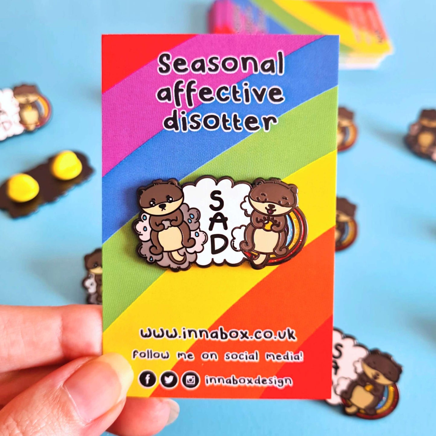 The seasonal affective disotter enamel pin on a rainbow backing card with black text above reading 'seasonal affective disotter' and below text of the innabox website and social handles being held over a blue background with other innabox pins. The enamel pin features two otters laying on a white cloud, on the left is a sad crying otter with raining grey clouds and on the right is a happy smiling otter with a rainbow. Raising awareness for SAD seasonal affective disorder.