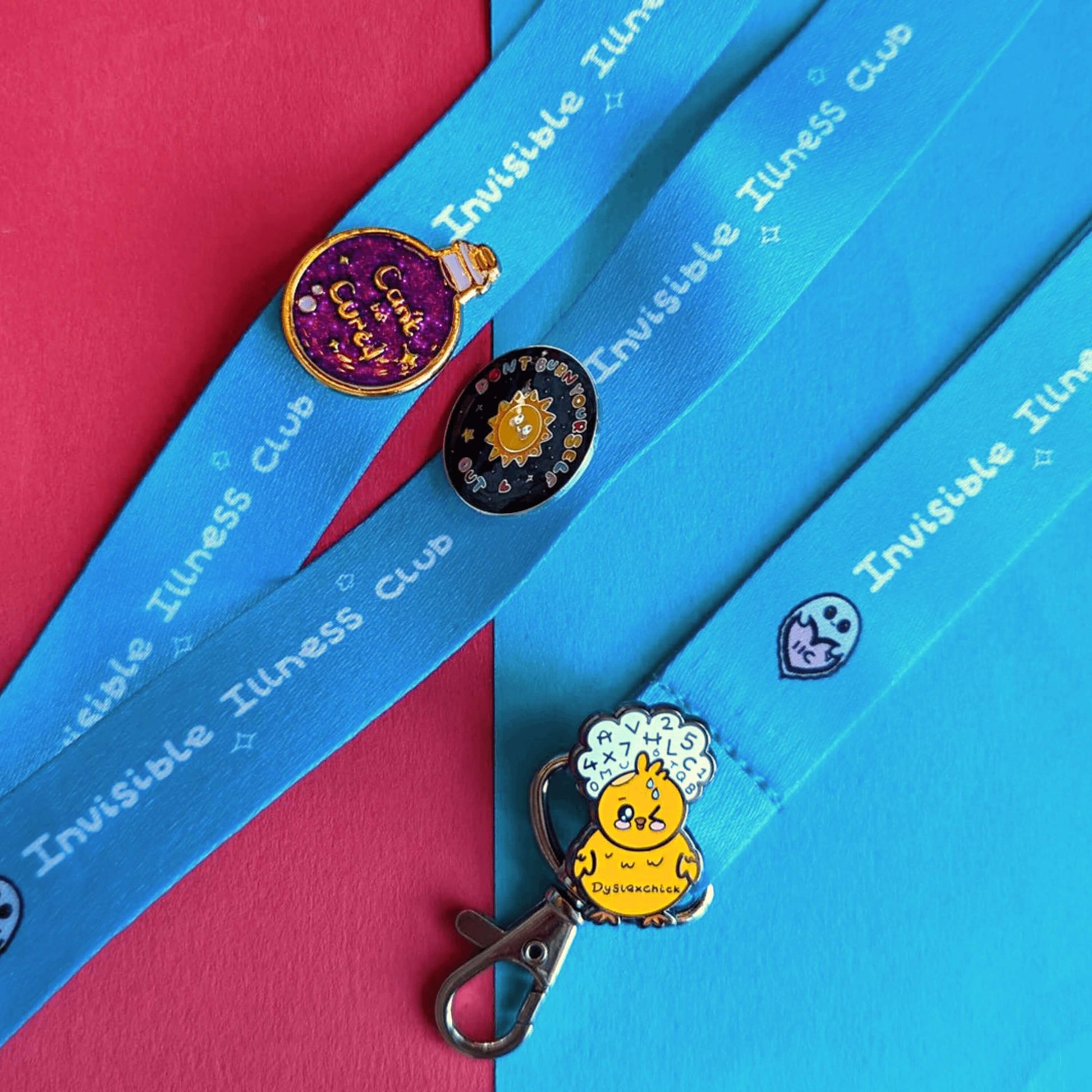 Dyslexia enamel pin of a yellow confused chick with numbers in a cloud above their head, Can't Be Cured Purple and Gold glitter Potion Bottle Enamel Pin and Don't Burn Yourself Out Sunshine enamel pin attached to Invisible Illness Club Lanyard. The background is blue and red.
