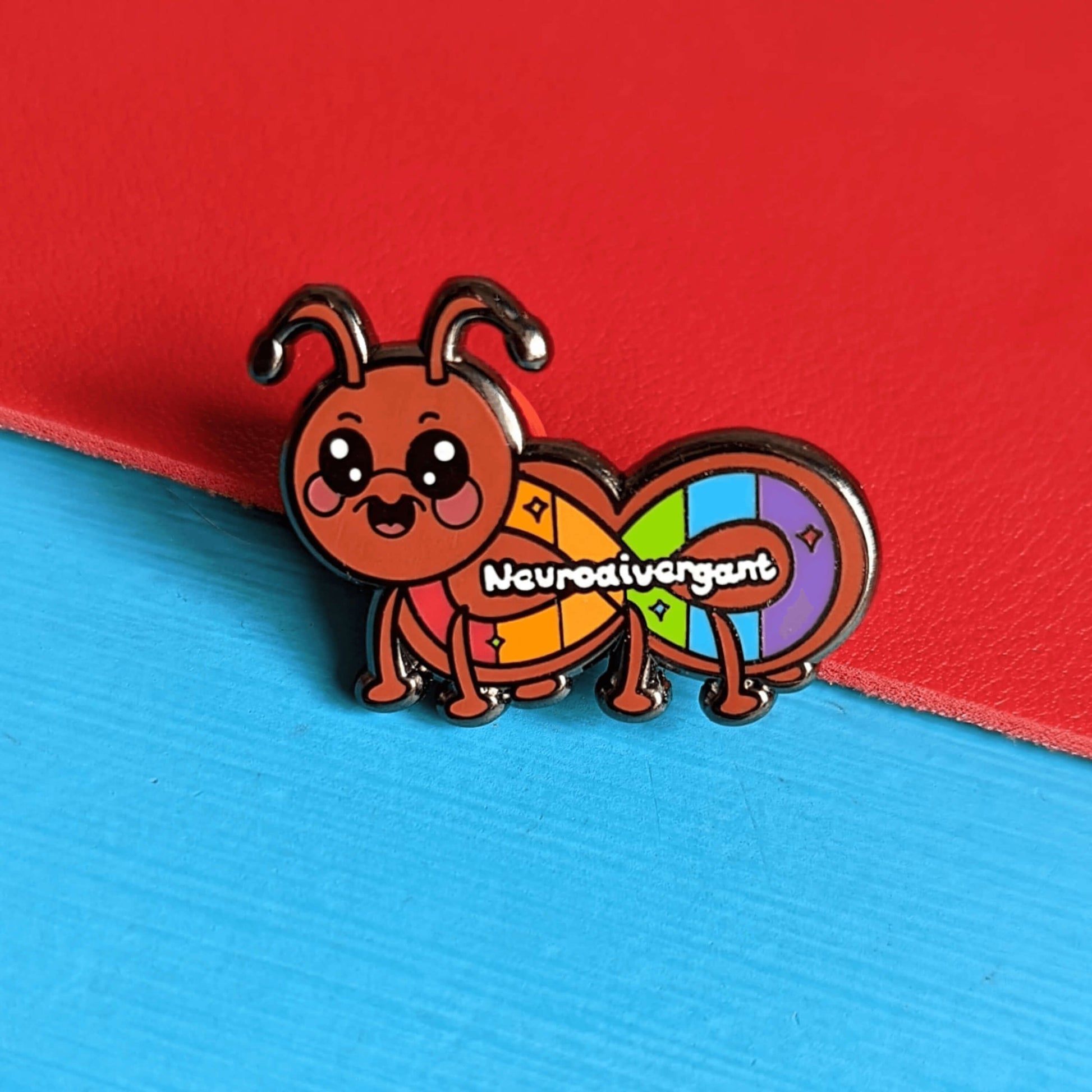 A cute brown ant enamel pin with wide sparkly eyes, rosy cheeks and antennae. The ant has it's mouth open in a smile and has the rainbow neurodivergent sign on it's body with 'neurodivergant' written in white across it. The enamel pin is shown on a red and blue background. Raising awareness for neurodivergence.