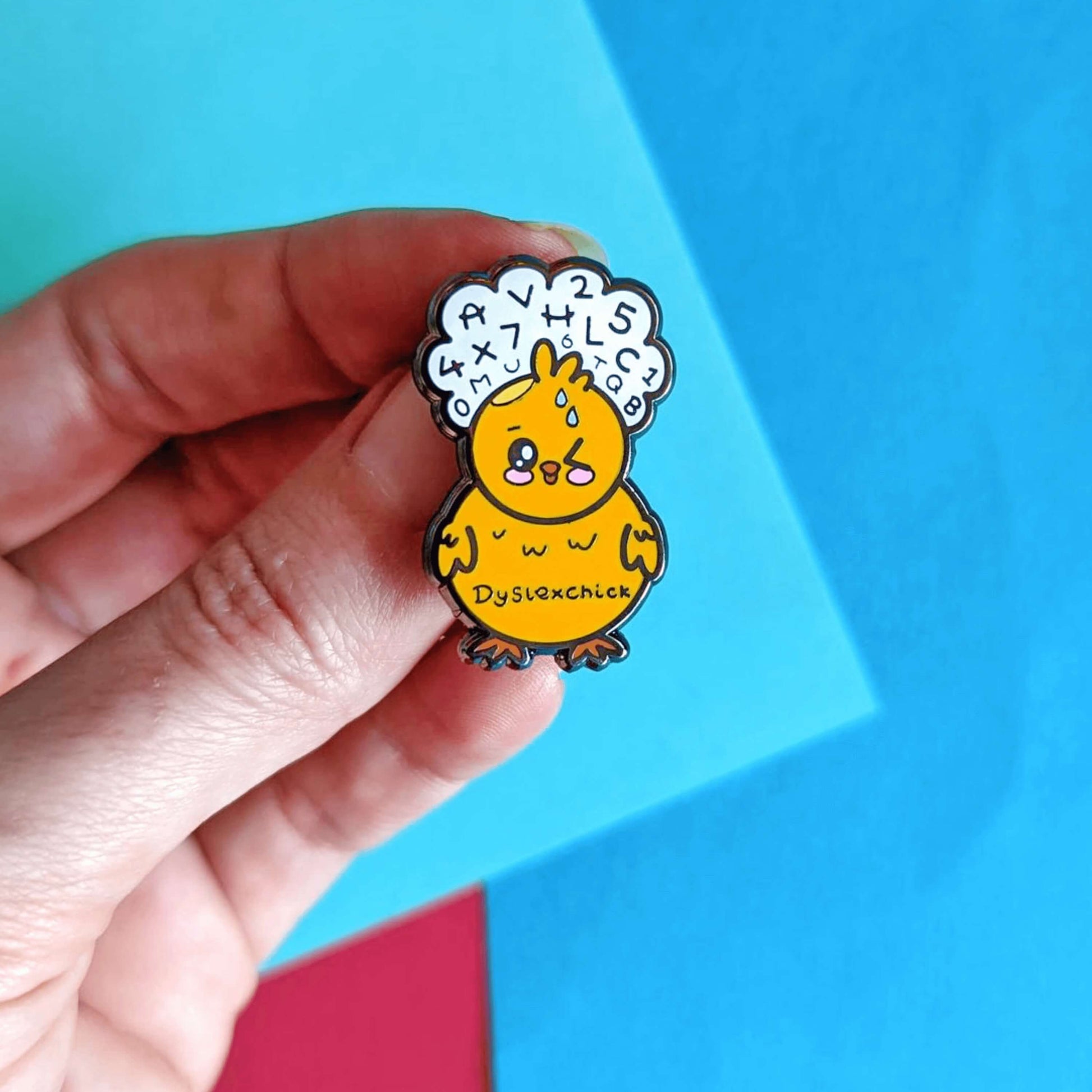 A cute yellow chick enamel pin with one eye closed shut and rosy cheeks. The chick has an orange beak and feet and a fluffy belly. There is a white cloud above the chicks head with various letters and numbers in. The chick also has sweat droplets on its forehead. 'Dyslexchick' is written across the chicks tummy in black. The pin is being held by a hand in front of a red and blue background. Design is raising awareness for dyslexia.