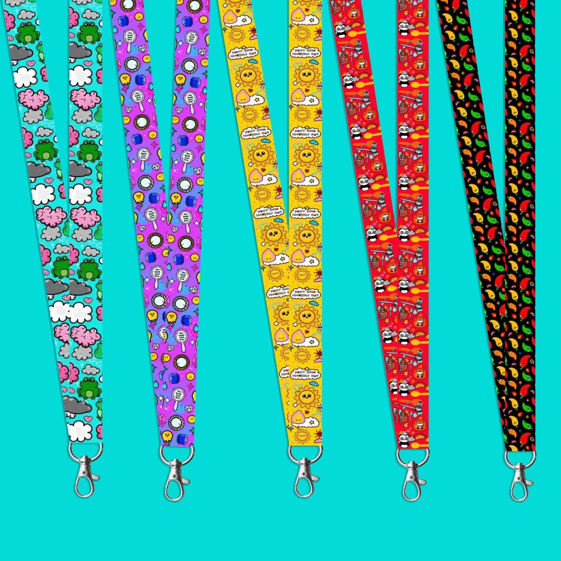 Brain Frog lanyard featuring various green frogs and pink, grey, white clouds with white sparkles, You Don't Look Sick lanyard of mirrors and gems with melting smiley faces, Burn Out lanyard of yellow, red sunshines, clouds and fires on yellow, Spoonie lanyard of various animals with spoons, fire and rainbows, and Neuro-Spicy lanyard of green, orange, red, yellow chilli peppers with rainbows and flames on a blue background.