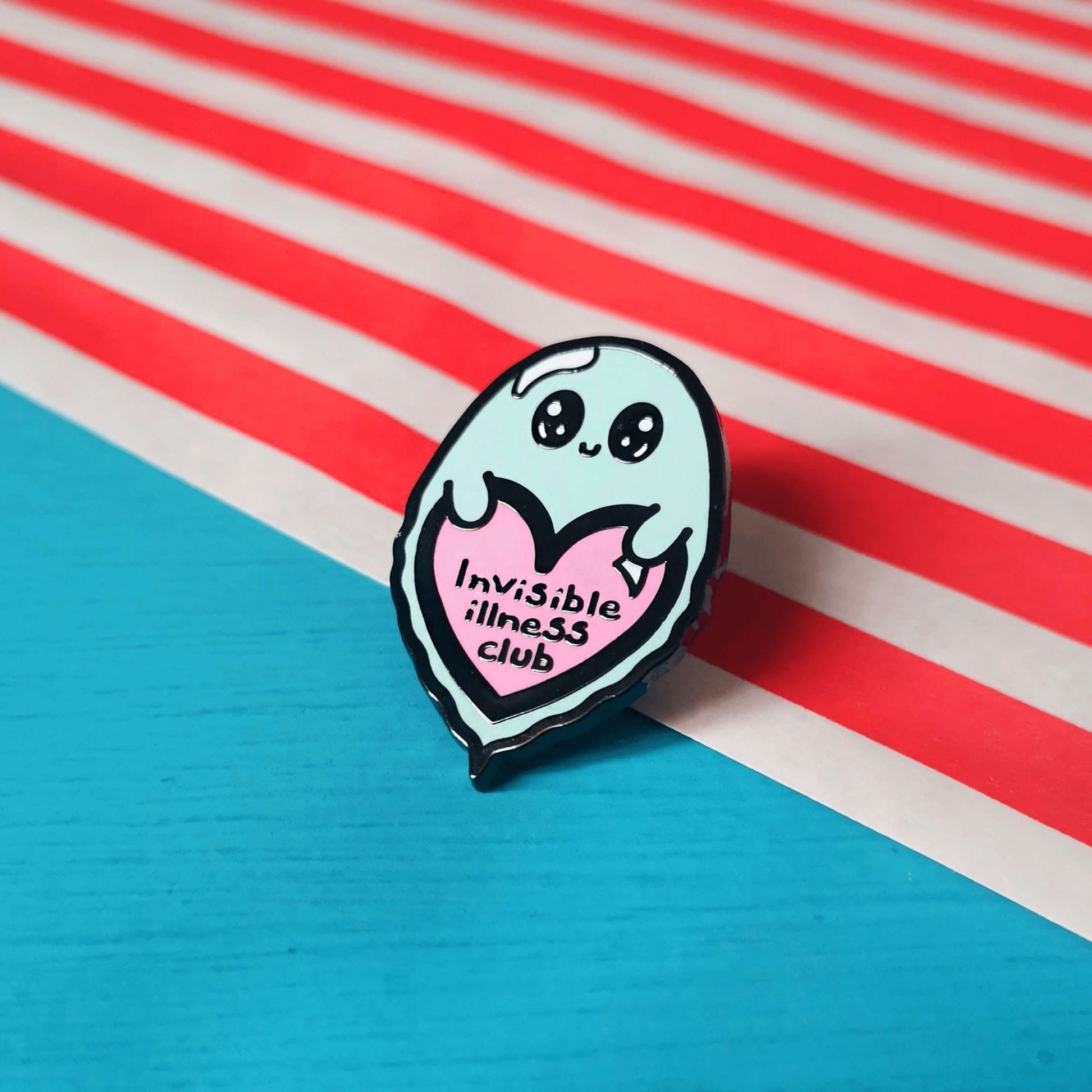 A cute blue ghost enamel pin with wide sparkly eyes and little smile holding a pink heart with it's little hands. 'Invisible illness club' is written in the heart in black. The enamel pin is on a red and white striped and blue background. Raising awareness for invisible illnesses.