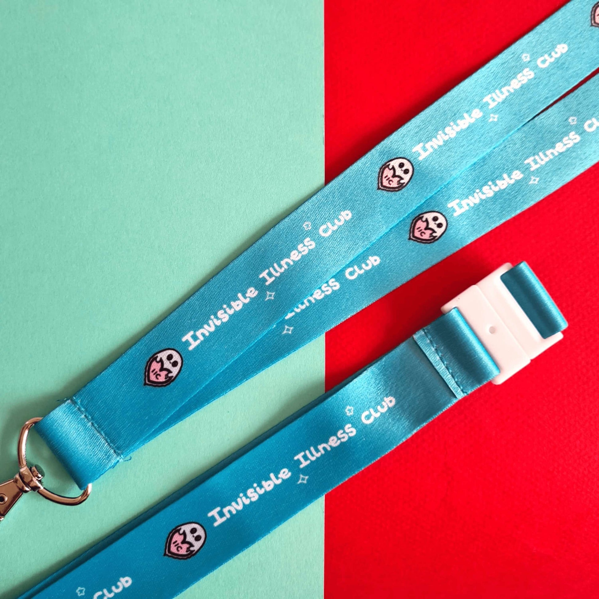 Pastel blue lanyard with 'Invisible Illness Club' written on it in white with Innabox ghost logo. The lanyard has a white clasp and silver lobster clip. Shown on a red and blue background. Raising awareness for invisible illnesses.