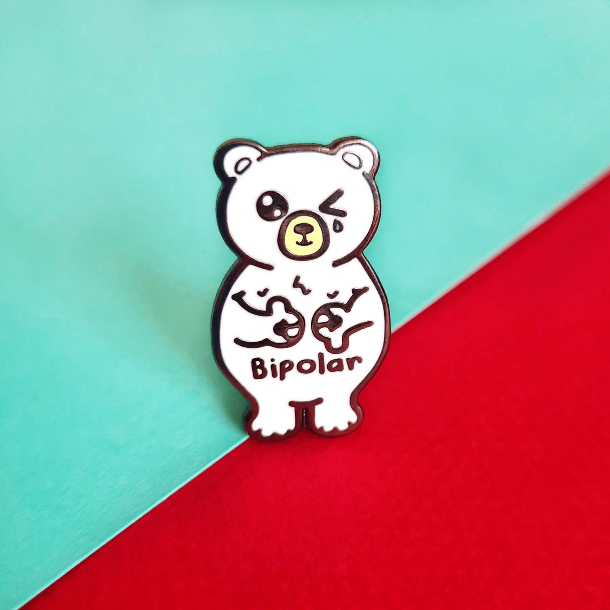 A white polar bear shaped enamel pin with one eye closed shut and a tear coming down its face. The polar bear has a fluffy tummy and has one thumb up and one thumb down. 'Bipolar' is written in black across its tummy. The background of the photo is red and blue.