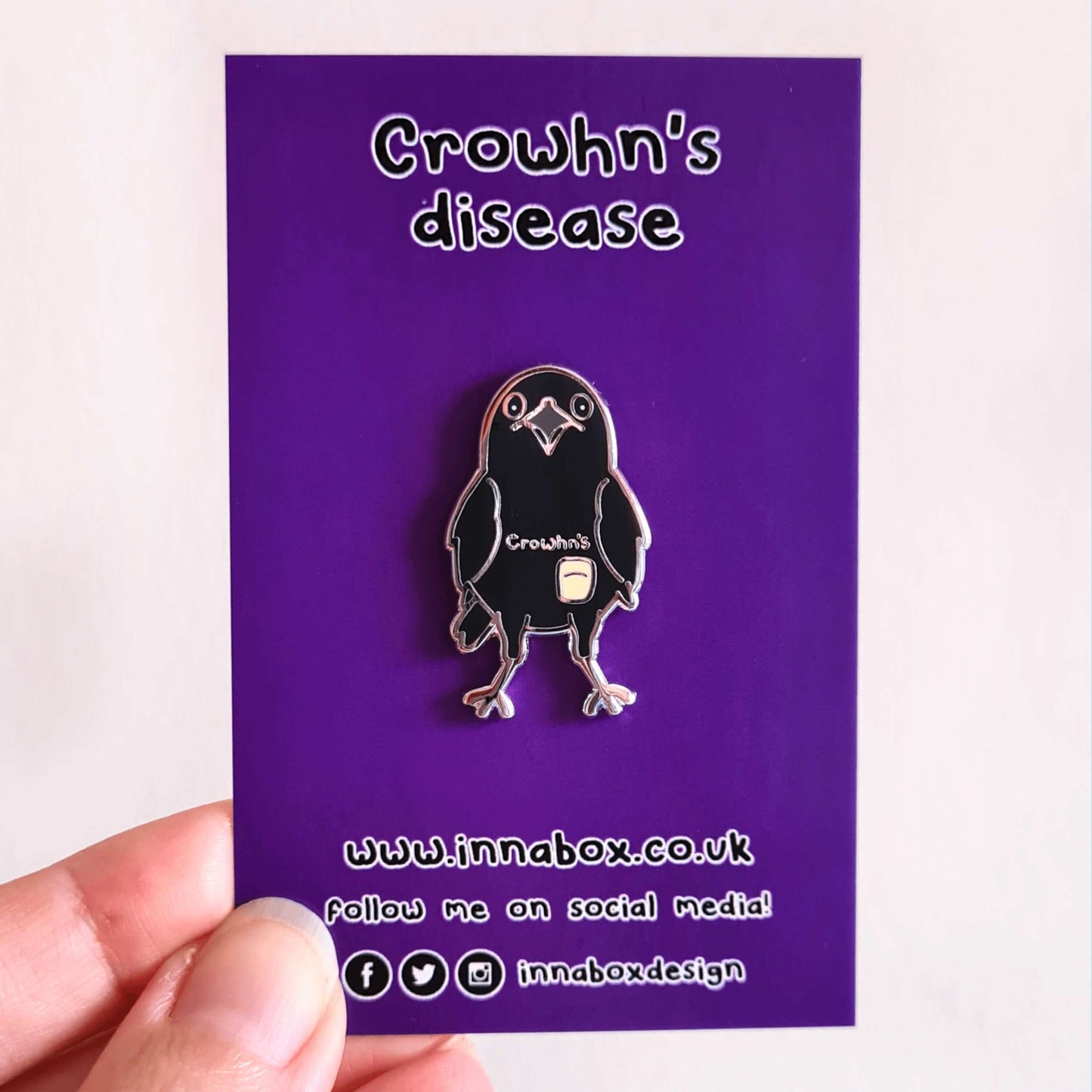 A black crow shaped enamel pin with silver beak, wings and feet. 'Crowhn's' is written in silver on the crow's tummy and there is a colostomy bag underneath the writing on the crow. The pin is attached to it's purple backing card with Innabox social media info at the bottom.