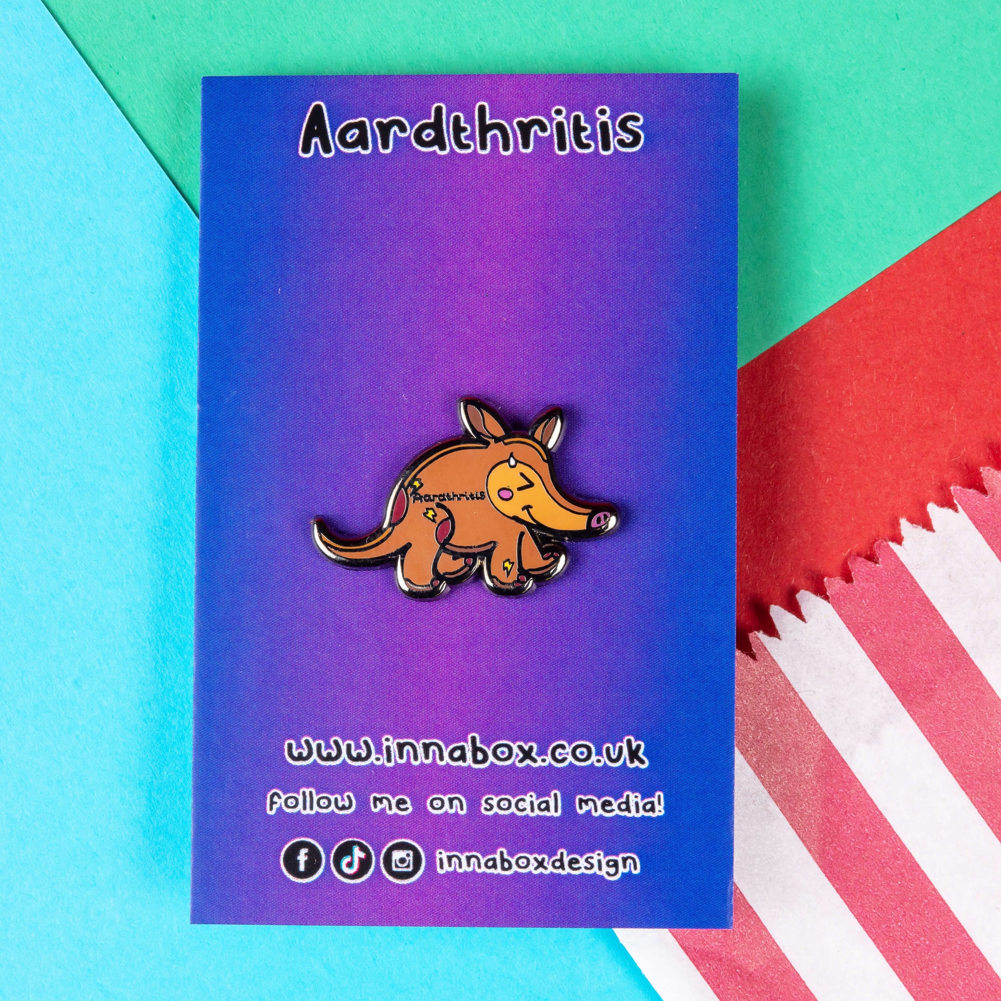 The Aardthritis Aardvark Enamel Pin - Arthritis on purple and blue gradient backing card with black text. The smiling sweating brown aardvark with pink cheeks and lightning bolts scattered across its body with black text reading 'aardthritis'. The hand drawn design is raising awareness for arthritis, Osteoarthritis and Rheumatoid arthritis.