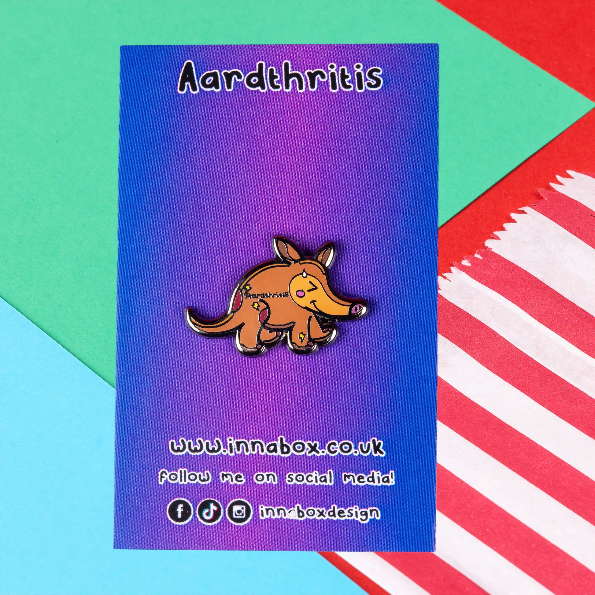 The Aardthritis Aardvark Enamel Pin - Arthritis on purple and blue gradient backing card with black text. The smiling sweating brown aardvark with pink cheeks and lightning bolts scattered across its body with black text reading 'aardthritis'. The hand drawn design is raising awareness for arthritis, Osteoarthritis and Rheumatoid arthritis.