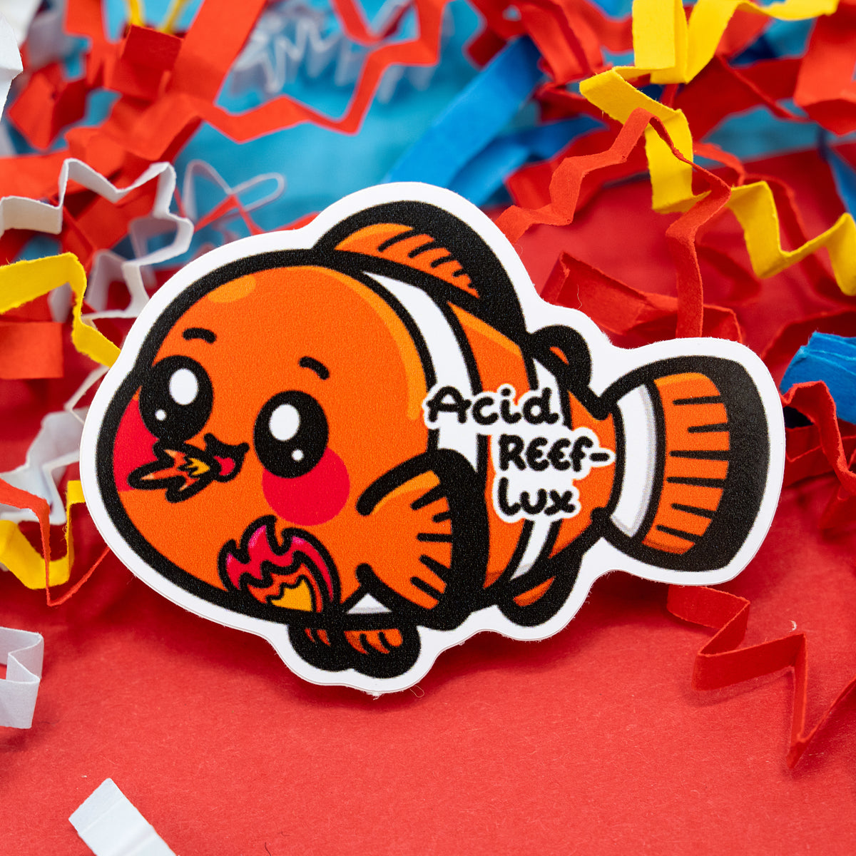 A cute vinyl sticker by Innabox featuring an orange clown fish pun design called “Acid Reef-Lux.” The fish has big eyes, rosy cheeks, and a fiery burp to represent acid reflux. Set against colourful shredded paper, this waterproof gloss vinyl sticker is about 5 cm tall and suitable for indoor and outdoor use.