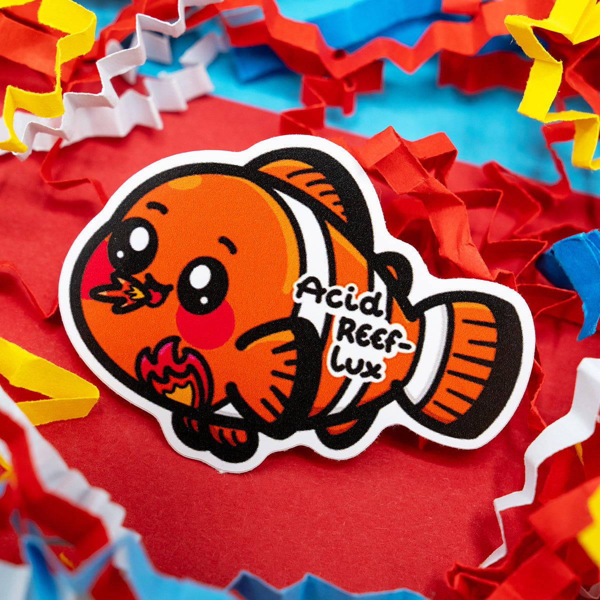 Close-up of the Innabox Acid Reef-Lux clown fish sticker, showing a bright orange fish character breathing a small flame in a fun pun on acid reflux. Surrounded by vibrant shredded paper, this waterproof gloss vinyl sticker is around 5 cm tall and perfect for indoor and outdoor use.