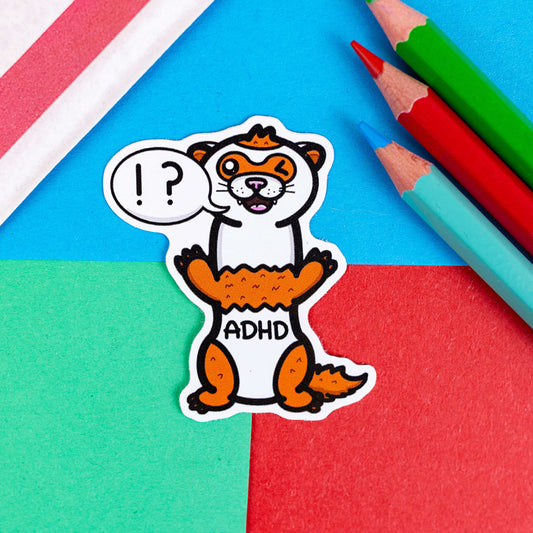 A red and white ADHD ferret sticker with it's mouth open in a smile and it's little teeth are showing. One of it's eyes are closed shut and it is standing on it's back legs. There is a speech-bubble from the ferret's mouth with '!?' inside it. The sticker is shown on a blue, red and green background with colouring pencils.