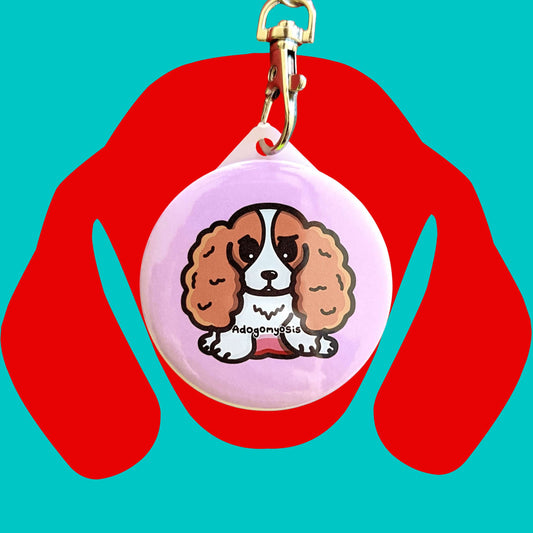 A sad spaniel dog with a red tummy and black text reading adogomyosis on a pink background plastic circular keychain hanging on a silver lobster clip. Adogomyosis Keyring - Adenomyosis is on a red and blue background. Raising awareness for adenomyosis.