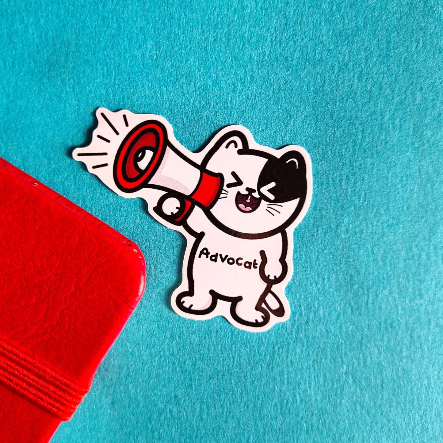 The Advocate Cat Sticker on a red and blue background. The black and white cat shape vinyl sticker is smiling shouting into a red and white megaphone with black text across its middle reading 'advocat'. The hand drawn design is raising awareness for mental health and disabilities.