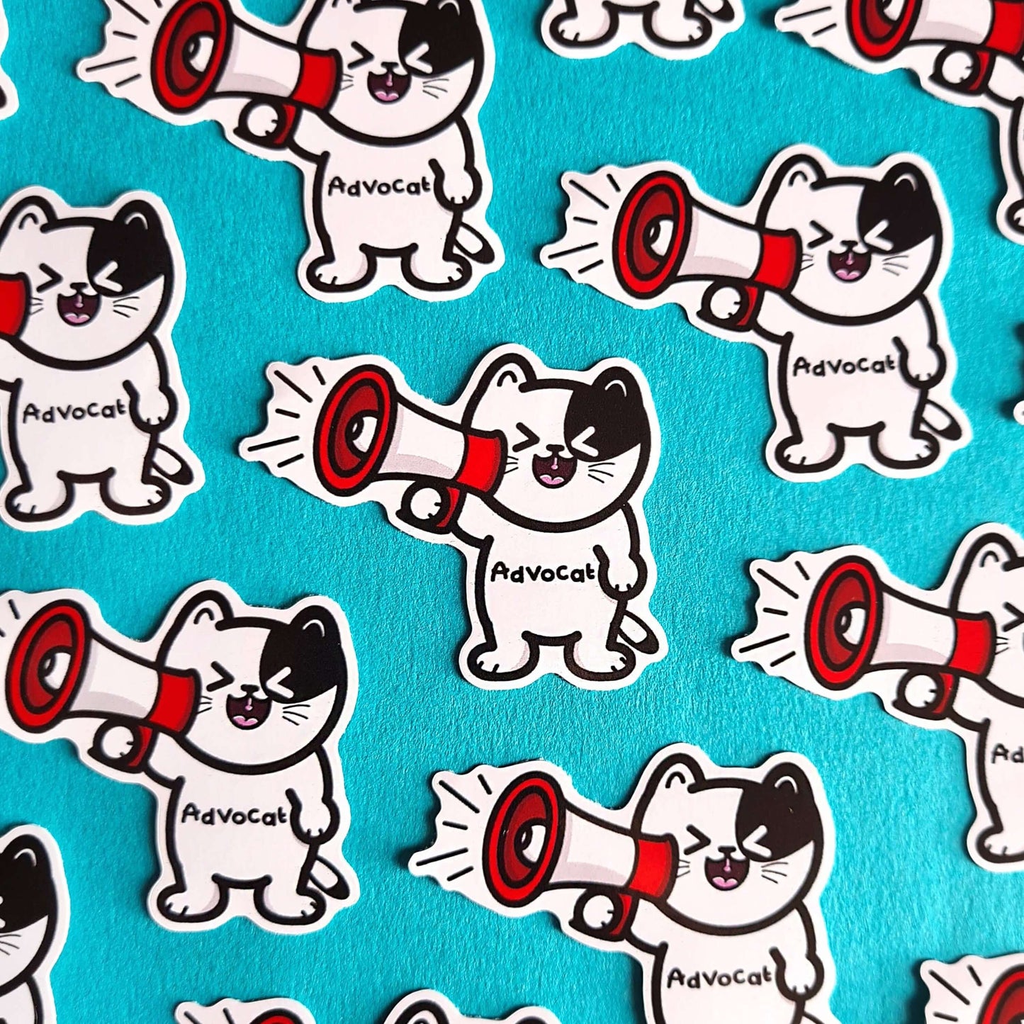 Multiple copies of the Advocate Cat Stickers on a blue background laid out in a repeating pattern. The black and white cat shape vinyl sticker is smiling shouting into a red and white megaphone with black text across its middle reading 'advocat'. The hand drawn design is raising awareness for mental health and disabilities.