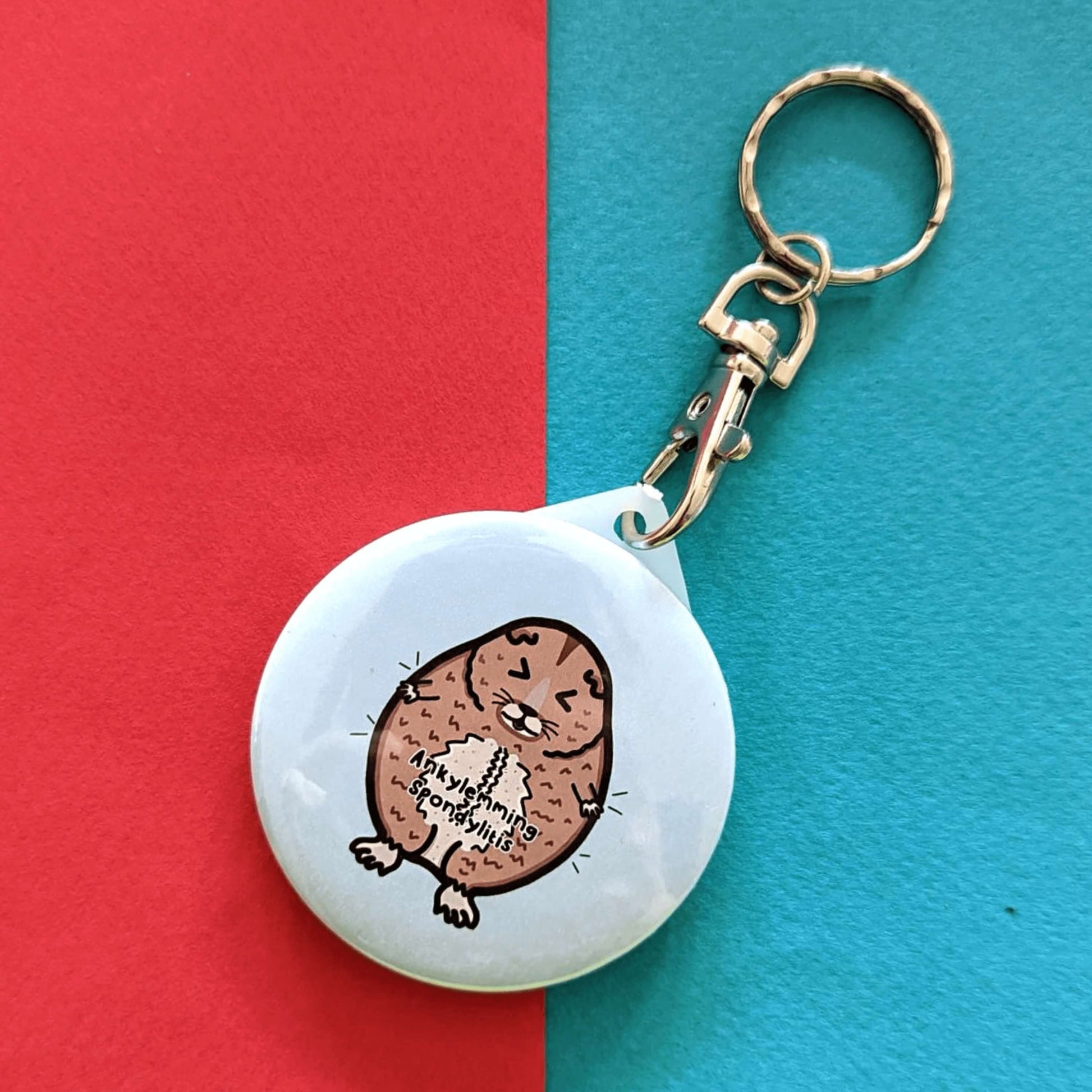 The Ankylemming - Ankylosing Spondylitis Keyring, a stressed lemming with its eyes scrunched shut on a white background circle keychain with the text ankylemming spondylitis being held up by a silver lobster clip on a blue and red background. Raising awareness for Ankylosing Spondylitis.