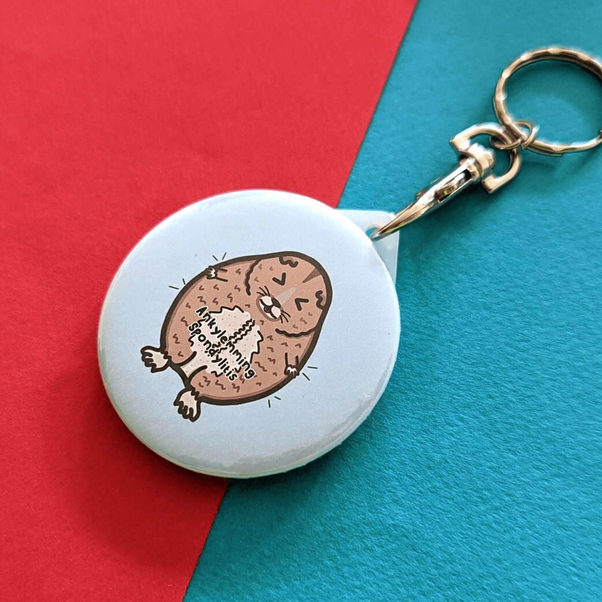 The Ankylemming - Ankylosing Spondylitis Keyring, a stressed lemming with its eyes scrunched shut on a white background circle keychain with the text ankylemming spondylitis being held up by a silver lobster clip on a blue and red background. Raising awareness for Ankylosing Spondylitis.